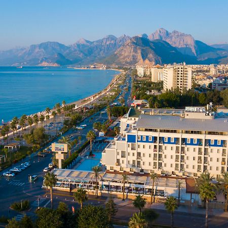 Sealife Family Resort Hotel Antalya Exterior foto The photo depicts a scenic coastal view, featuring a beachfront area lined with palm trees. In the foreground, there is a hotel or resort with a pool and outdoor seating. The road runs parallel to the beach, with some vehicles visible. In the backgro