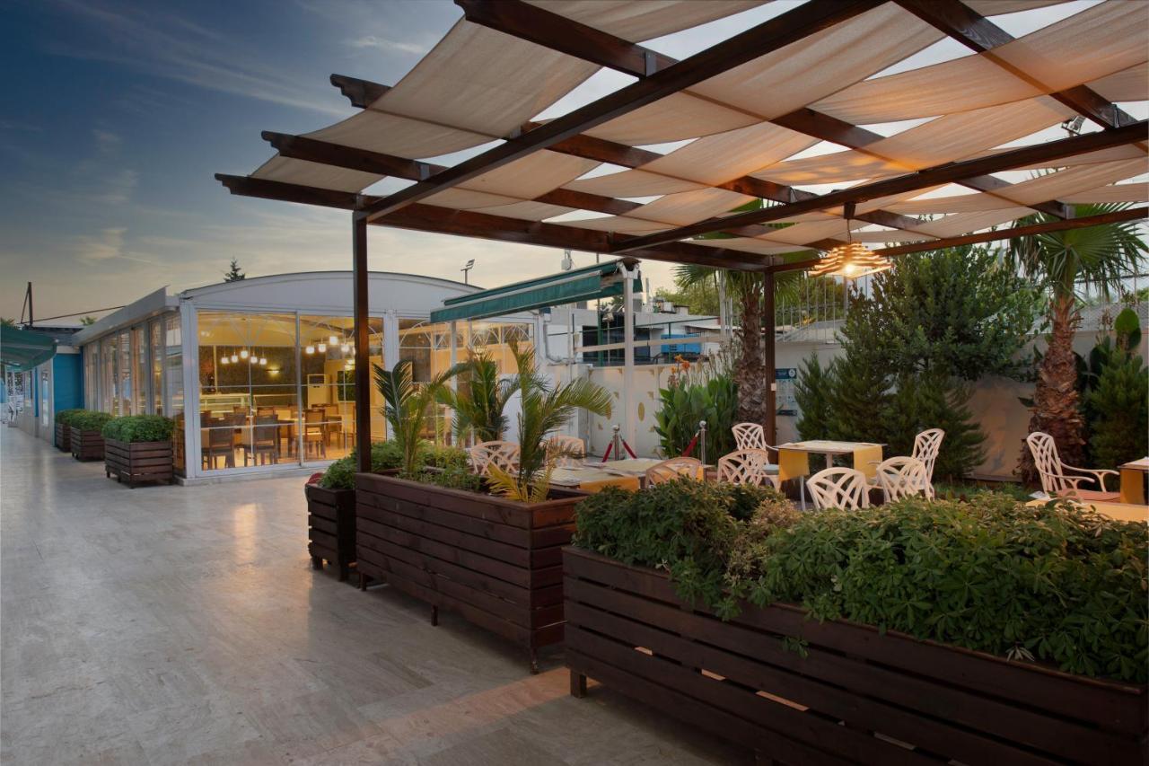 Sealife Family Resort Hotel Antalya Exterior foto The photo depicts an outdoor space, likely a café or restaurant terrace. It features a wooden structure with a grid-like canopy that provides shade, and several white chairs with decorative designs are arranged around small tables. There are large pl