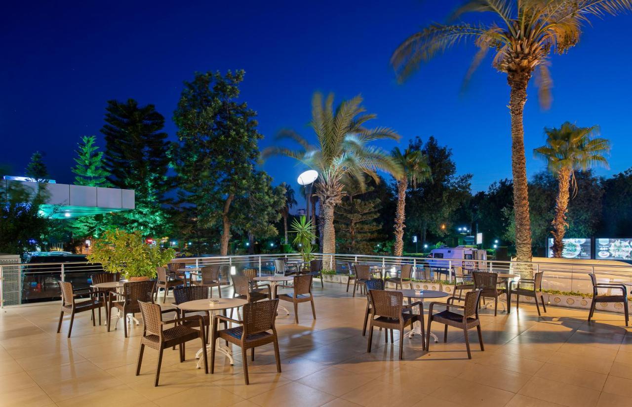 Sealife Family Resort Hotel Antalya Exterior foto The photo depicts an outdoor dining area, likely part of a restaurant or café, set in the evening. There are several tables with chairs arranged across a tiled patio. The space is surrounded by tall palm trees and greenery, creating a tropical ambian