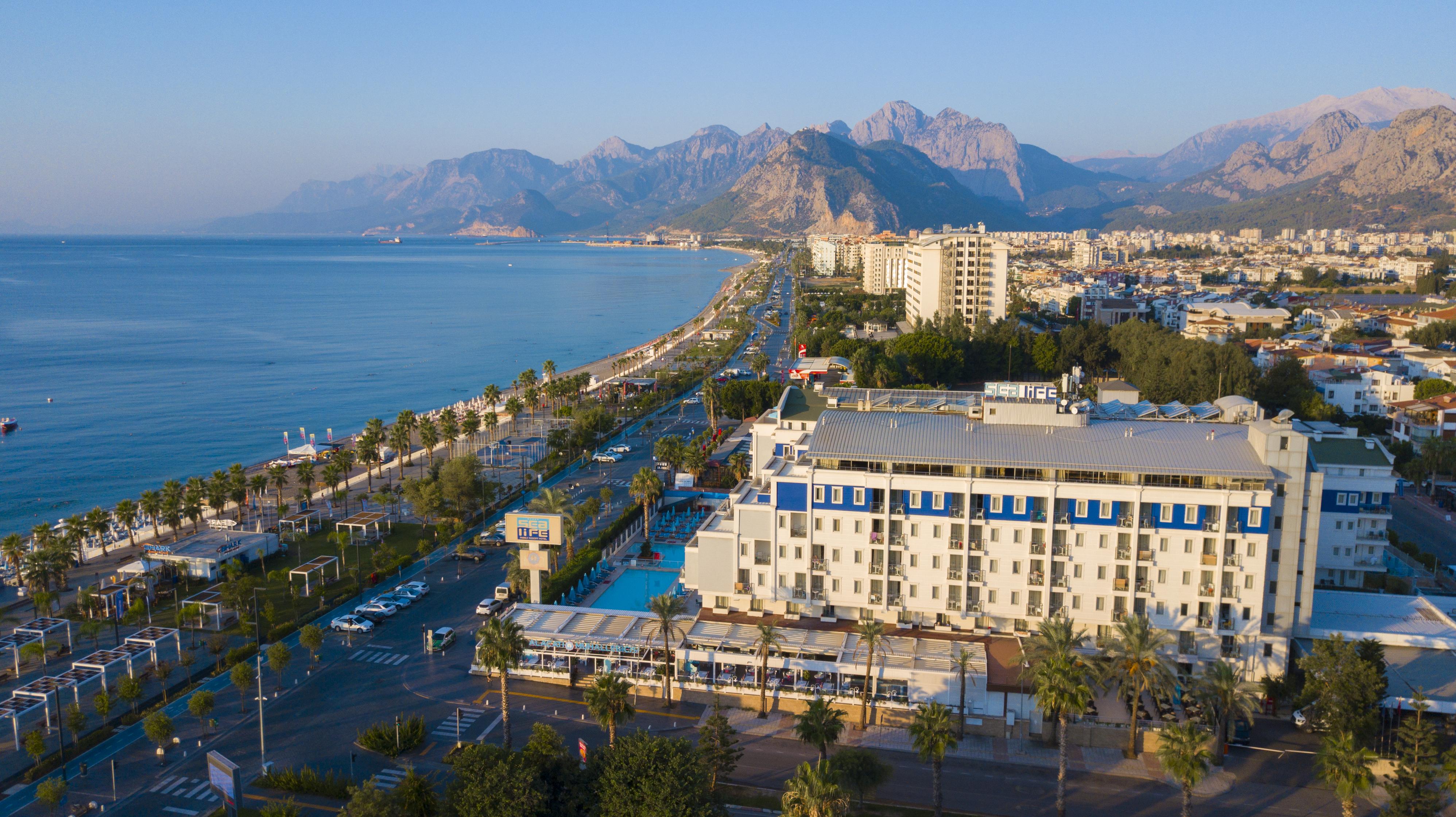 Sealife Family Resort Hotel Antalya Exterior foto The photo depicts a scenic coastal view, featuring a beachfront area lined with palm trees. In the foreground, there is a hotel or resort with a pool and outdoor seating. The road runs parallel to the beach, with some vehicles visible. In the backgro