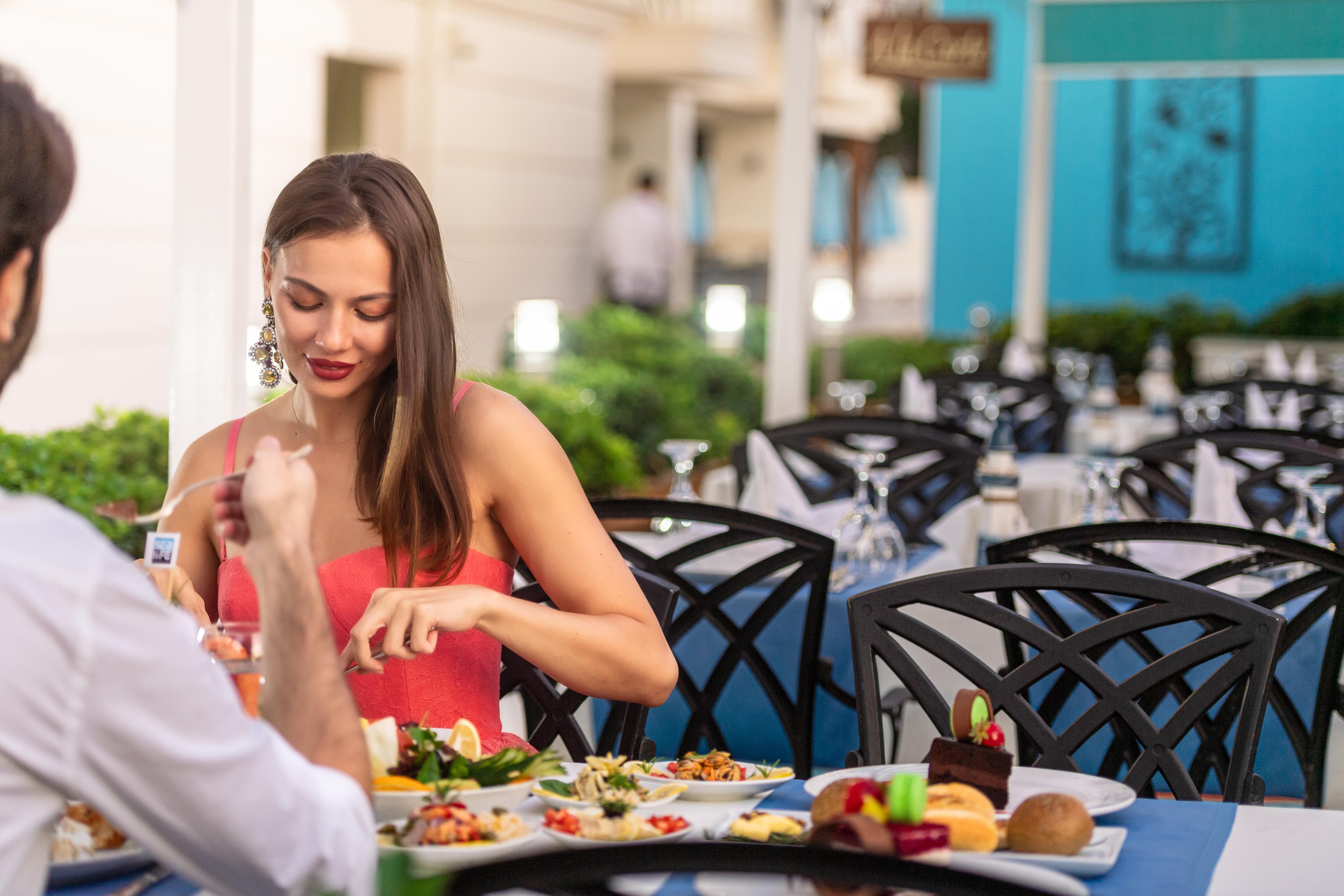Sealife Family Resort Hotel Antalya Exterior foto The photo shows a dining scene at a restaurant. A woman in a red dress is seated at a table, interacting with her meal. She appears to be taking time to enjoy her food. The table is set with various dishes, including salads and desserts. The backgrou