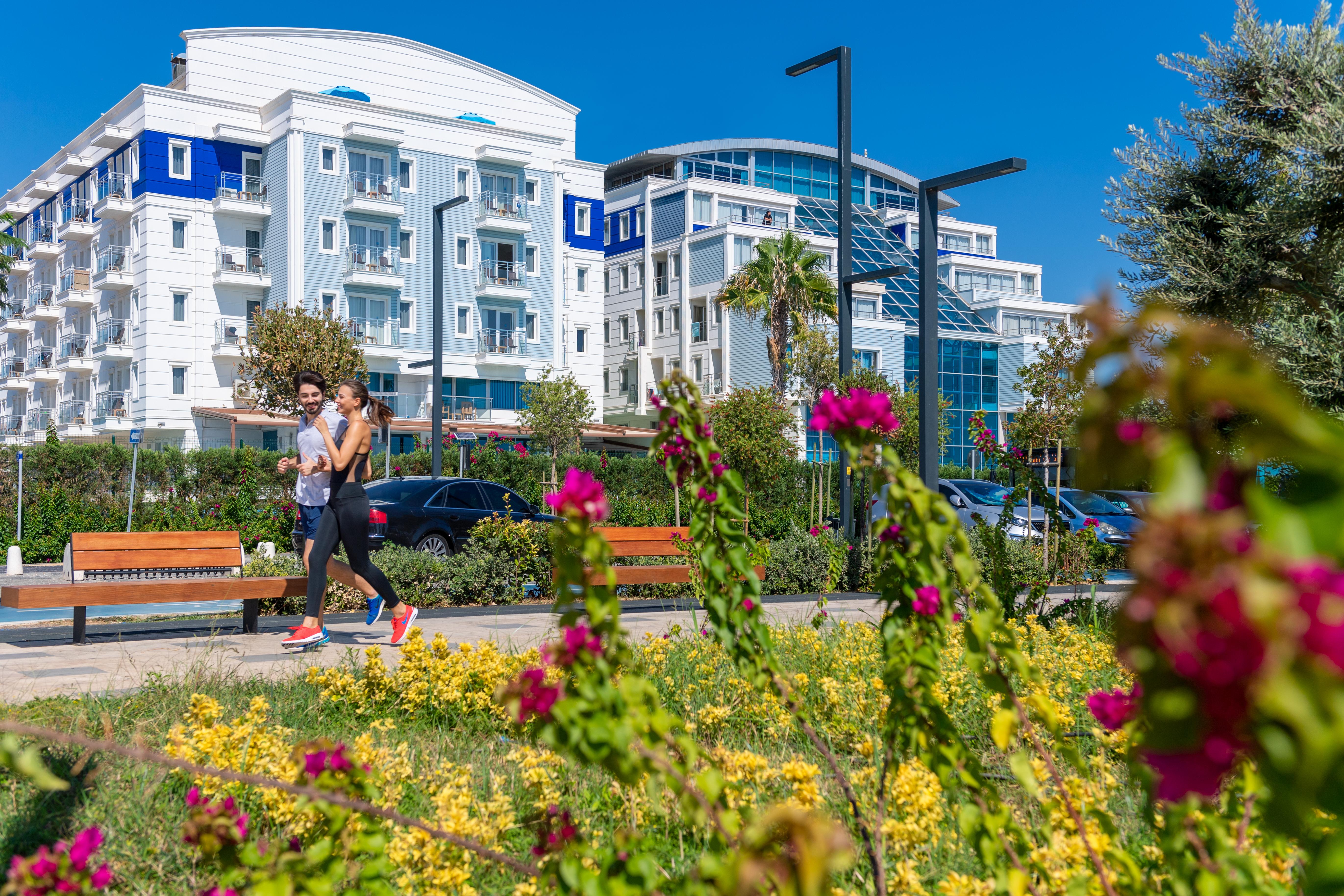 Sealife Family Resort Hotel Antalya Exterior foto The photo depicts a sunny outdoor scene featuring two individuals walking along a path. They are surrounded by a vibrant landscape filled with yellow flowers and pink bougainvillea. In the background, there are modern buildings with balconies, some h