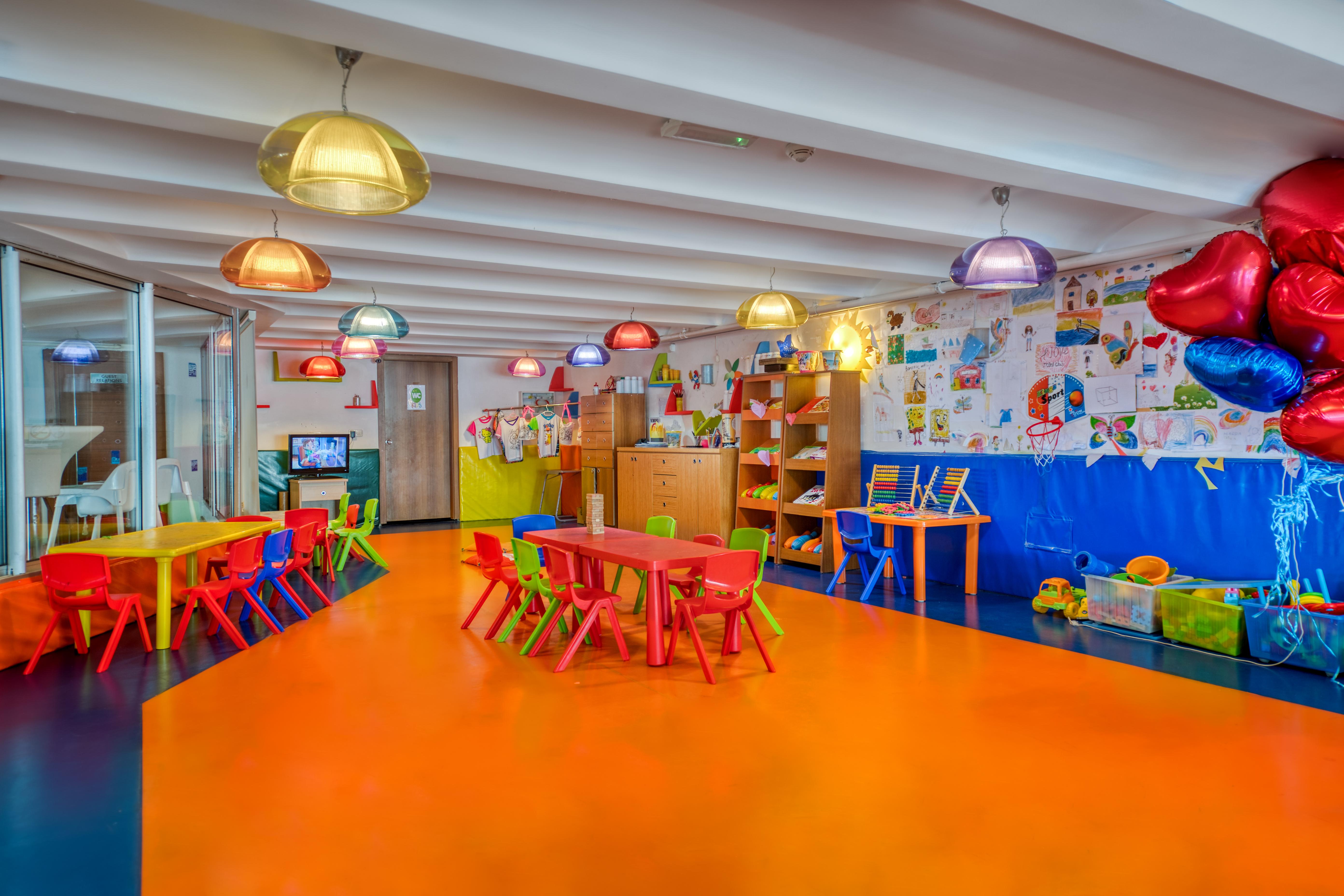 Sealife Family Resort Hotel Antalya Exterior foto The photo shows a vibrant and colorful kindergarten classroom. The flooring is a bright orange, contributing to the cheerful atmosphere. Various tables and chairs are arranged throughout the space, with some in yellow and red. Colorful pendant lights