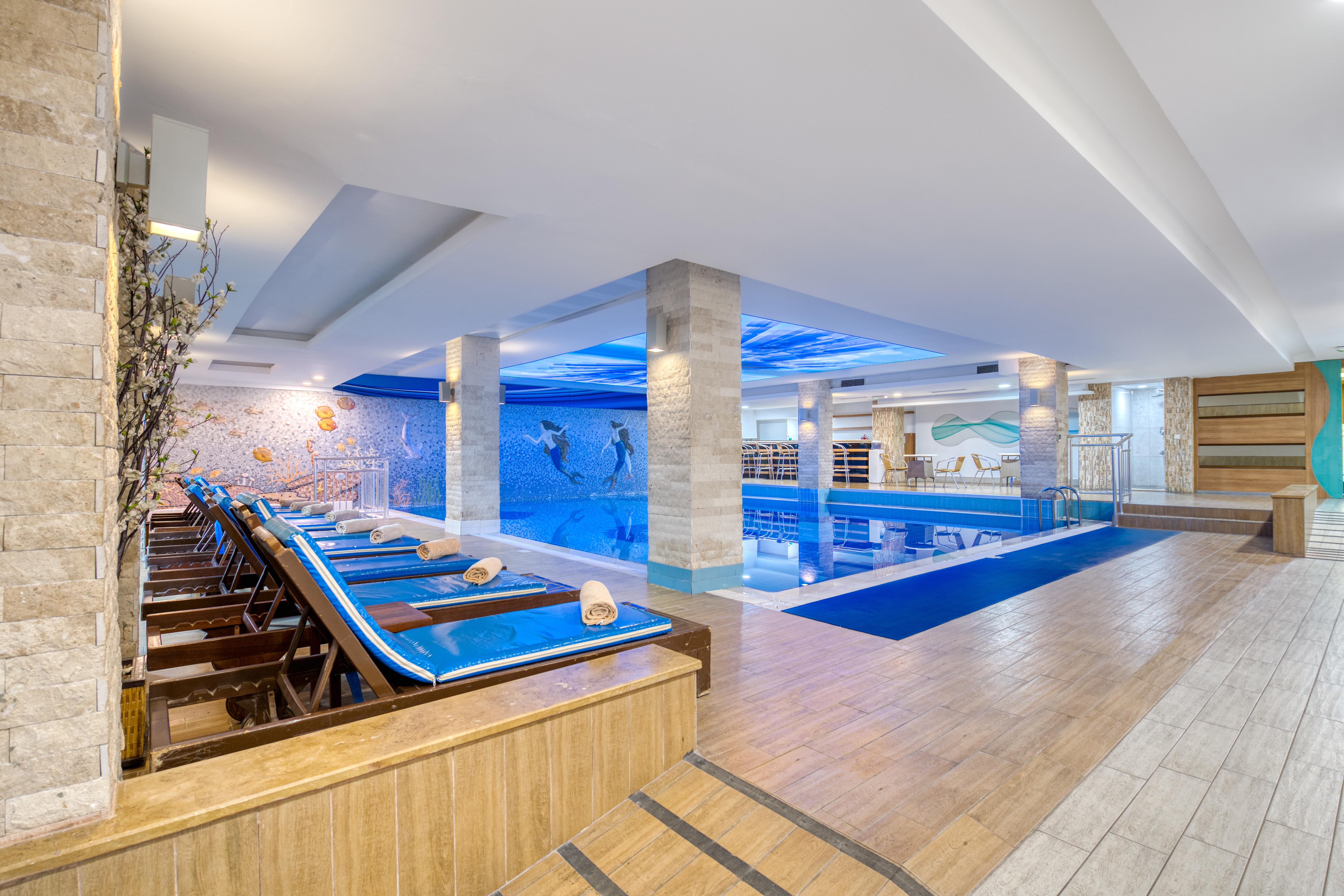 Sealife Family Resort Hotel Antalya Exterior foto The image shows an indoor swimming pool area. The pool is illuminated with soft blue lighting, creating a relaxing atmosphere. Alongside the pool, there are several lounge chairs arranged, some of which have rolled towels placed on them. The flooring