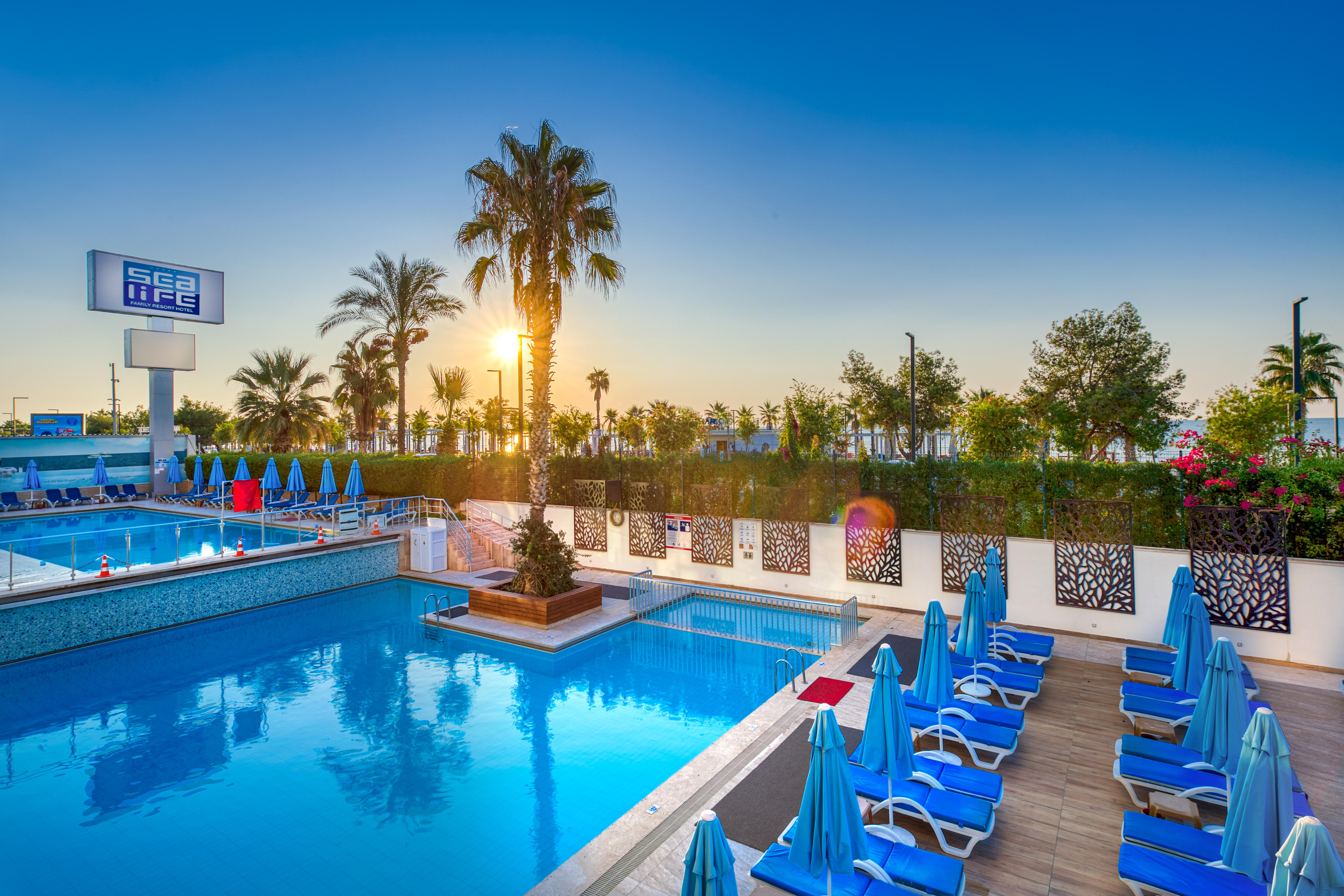 Sealife Family Resort Hotel Antalya Exterior foto The photo depicts a serene outdoor swimming pool area, illuminated by the warm light of a setting sun. There are several blue lounge chairs arranged around the pool, with palm trees and lush greenery in the background. The sky is clear, showcasing a 