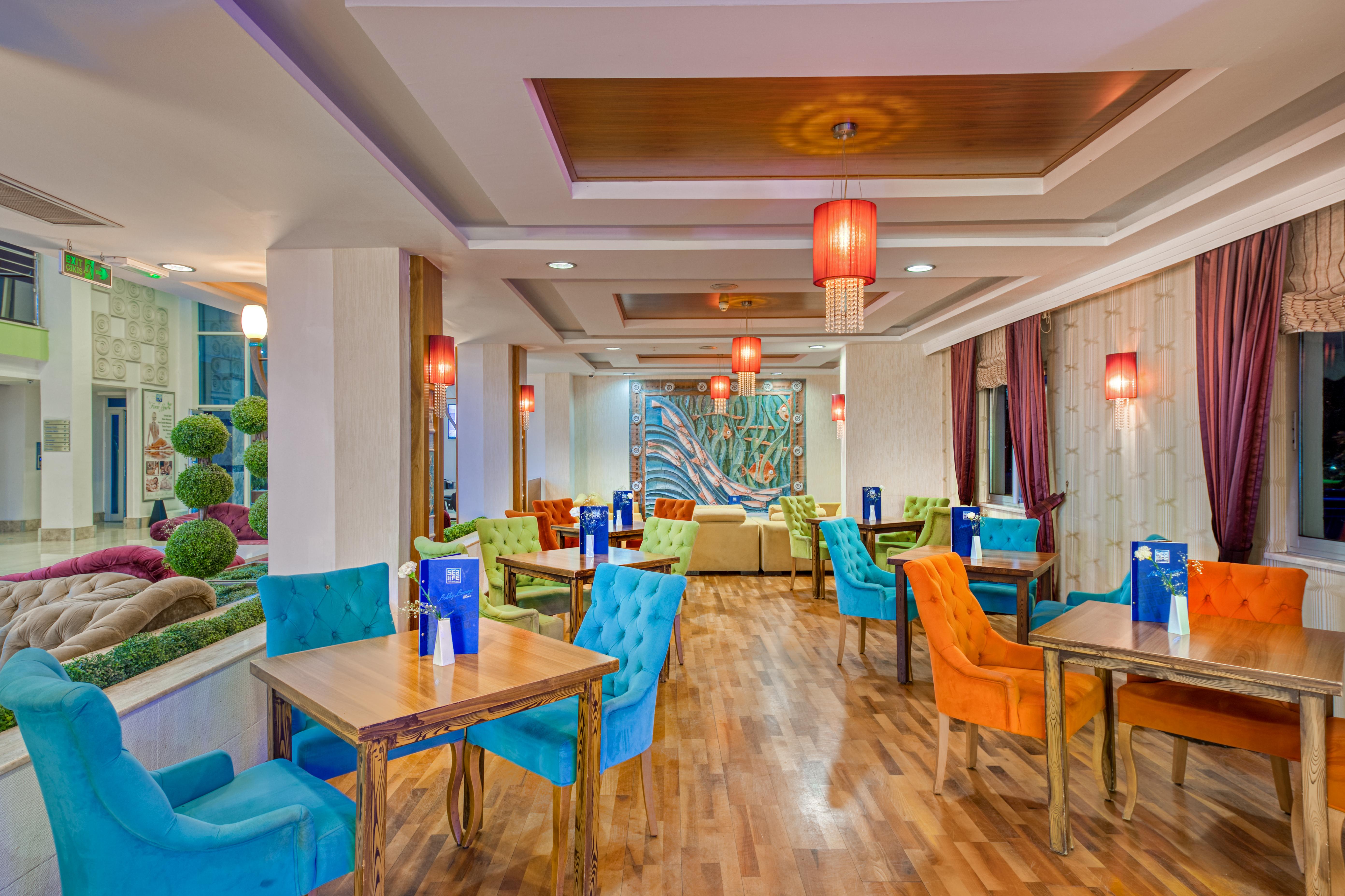 Sealife Family Resort Hotel Antalya Exterior foto The photo shows a stylish dining area with a contemporary design. The space features a combination of colorful chairs in blue and orange, and wooden tables arranged throughout. The walls have a warm, neutral tone and are complemented by decorative el