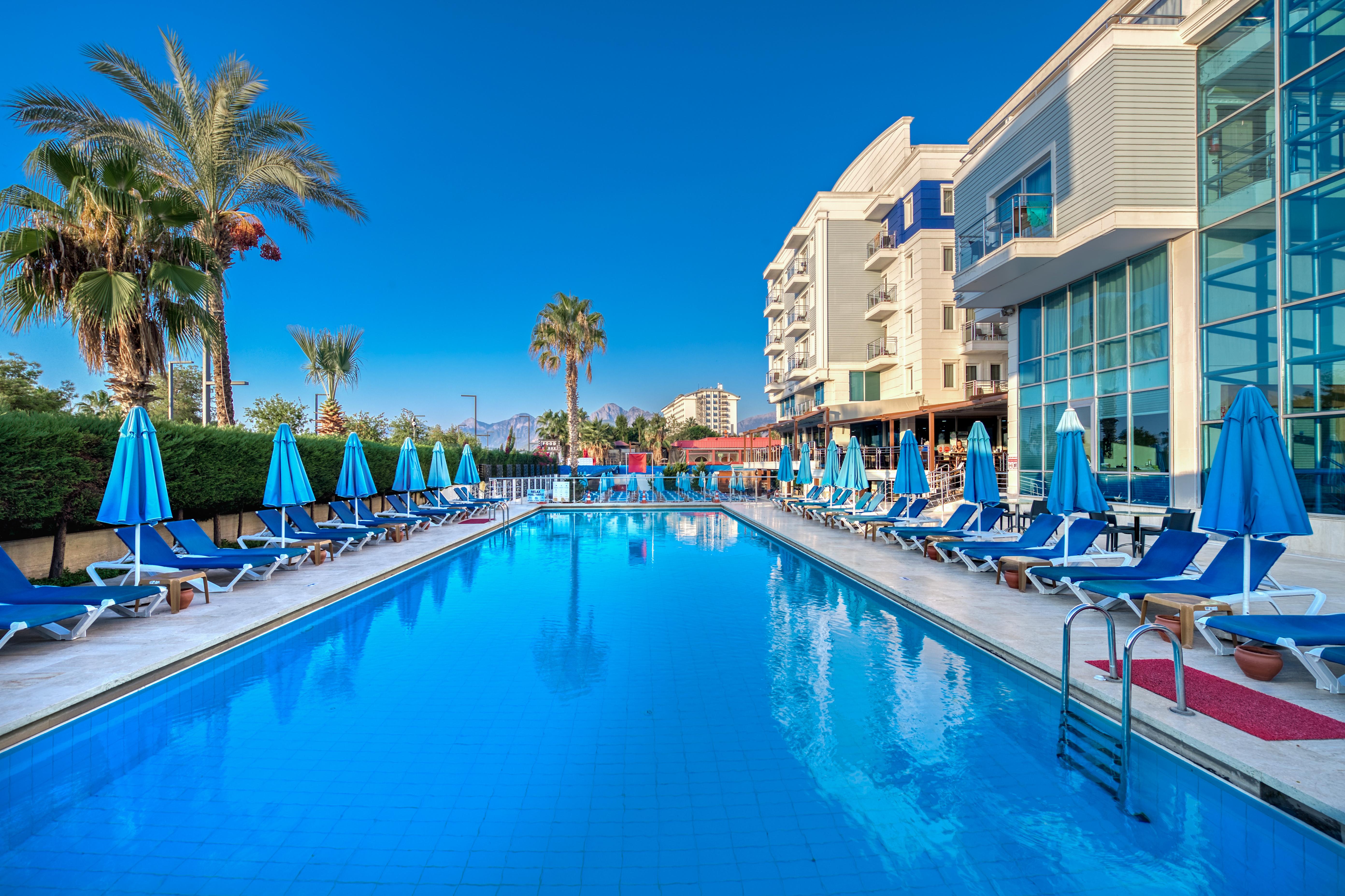 Sealife Family Resort Hotel Antalya Exterior foto The photo depicts a serene poolside scene at a hotel or resort. In the foreground, a large blue swimming pool reflects the clear sky, with comfortable lounge chairs arranged neatly around it under blue umbrellas. Lush palm trees add to the tropical a